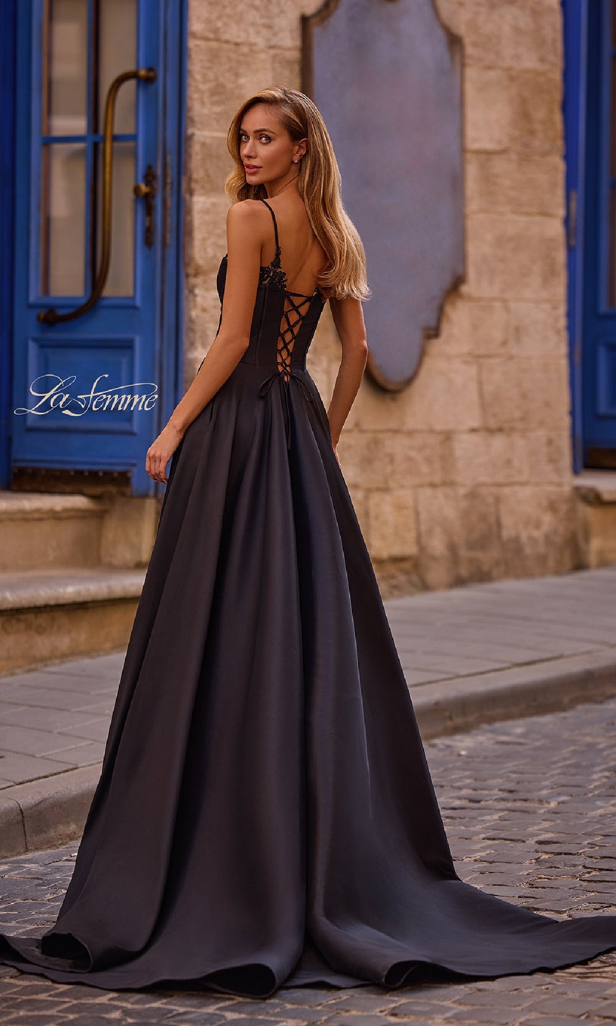 A woman wearing the La Femme 32878 Lace-Up Long Mikado Prom Dress stands on a cobblestone street near a stone building with blue doors and windows.